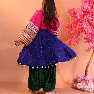 Puede incluir: Un atuendo tradicional vibrante, con una parte superior rosa con mangas bordadas ornamentadas, una falda azul fluida adornada con puntos blancos y pantalones verdes estampados. El conjunto se completa con zapatos a juego. El conjunto est&aacute; sobre un fondo melocot&oacute;n.