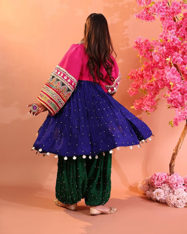 Puede incluir: Un atuendo tradicional vibrante, con una parte superior rosa con mangas bordadas ornamentadas, una falda azul fluida adornada con puntos blancos y pantalones verdes estampados. El conjunto se completa con zapatos a juego. El conjunto est&aacute; sobre un fondo melocot&oacute;n.