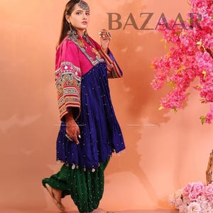 Puede incluir: Una mujer con un atuendo tradicional con una parte superior rosa, un vestido azul y pantalones verdes. El atuendo presenta intrincados bordados y adornos. El fondo es melocot&oacute;n con flores rosas y el texto "KOCHAI BAZAAR".