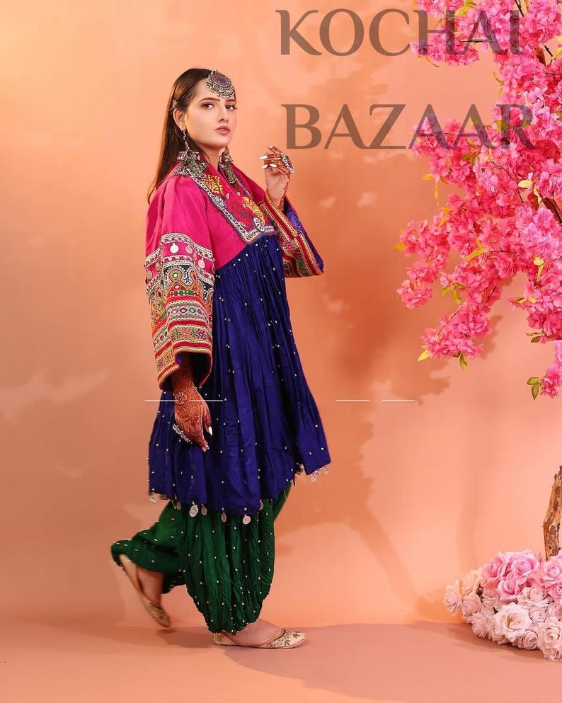Puede incluir: Una mujer con un atuendo tradicional con una parte superior rosa, un vestido azul y pantalones verdes. El atuendo presenta intrincados bordados y adornos. El fondo es melocot&oacute;n con flores rosas y el texto "KOCHAI BAZAAR".