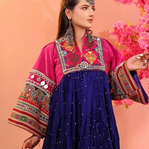 Puede incluir: Un vestido afgano tradicional en colores vibrantes. El vestido presenta un corpi&ntilde;o y mangas magenta con intrincados bordados y adornos. La falda es azul oscuro con peque&ntilde;os acentos dispersos. La modelo lleva pendientes y una diadema plateados.
