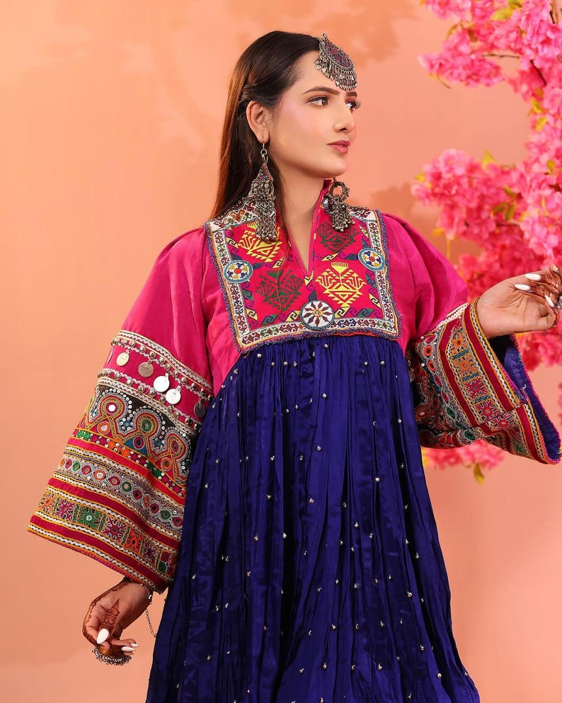 Puede incluir: Un vestido afgano tradicional en colores vibrantes. El vestido presenta un corpi&ntilde;o y mangas magenta con intrincados bordados y adornos. La falda es azul oscuro con peque&ntilde;os acentos dispersos. La modelo lleva pendientes y una diadema plateados.