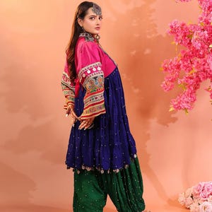 Puede incluir: Un atuendo tradicional con una parte superior rosa con mangas bordadas, un vestido azul marino y pantalones verdes. El conjunto incluye joyas de plata y se presenta sobre un fondo melocot&oacute;n, con flores rosas.