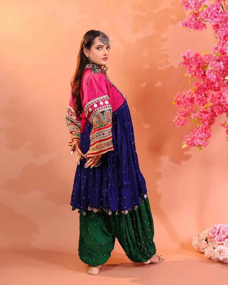 Puede incluir: Un atuendo tradicional con una parte superior rosa con mangas bordadas, un vestido azul marino y pantalones verdes. El conjunto incluye joyas de plata y se presenta sobre un fondo melocot&oacute;n, con flores rosas.