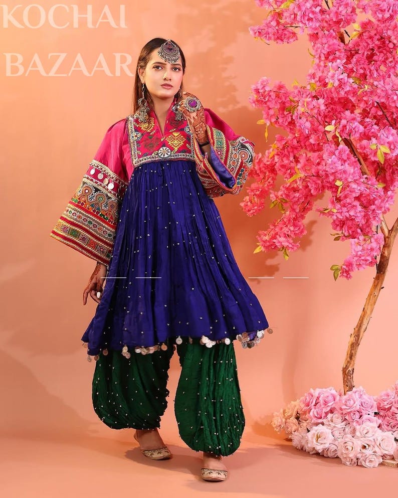 Puede incluir: Un vestido afgano tradicional con una parte superior rosa, una falda azul y pantalones verdes. El vestido presenta intrincados bordados y adornos. Las mangas son largas y acampanadas, y los pantalones son holgados. El texto "KOCHAI BAZAAR" es visible en la parte superior.