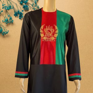 May include: Black long-sleeved tunic with a vertical tricolour design of black, red, and green. The red section features a gold emblem. The sleeves have red and green trim. The tunic is displayed on a mannequin.