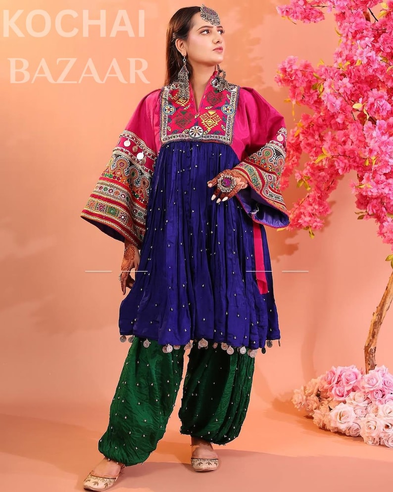 Puede incluir: Un vibrante vestido afgano tradicional con corpi&ntilde;o rosa, falda azul y pantal&oacute;n verde. El vestido presenta intrincados bordados y adornos. La imagen incluye el texto "KOCHAI BAZAAR".