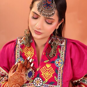 Puede incluir: Una mujer con joyer&iacute;a tradicional afgana, incluyendo una diadema plateada con detalles rojos, azules y plateados, y aretes a juego. Tambi&eacute;n lleva un vestido rosa con intrincados bordados y un gran anillo con una piedra azul.