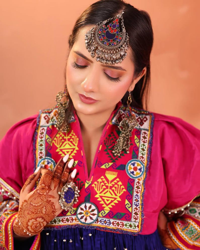 Puede incluir: Una mujer con joyer&iacute;a tradicional afgana, incluyendo una diadema plateada con detalles rojos, azules y plateados, y aretes a juego. Tambi&eacute;n lleva un vestido rosa con intrincados bordados y un gran anillo con una piedra azul.