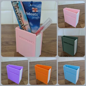 May include: A collection of rectangular plastic holders for bookmarks, each featuring the word BOOKMARKS embossed on the front. The holders are available in pink, dark green, purple, orange, and light blue, all with a white interior section.