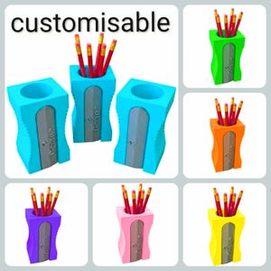 May include: A collection of pencil holders shaped like large pencil sharpeners in various bright colours including blue, green, orange, purple, pink, and yellow. Each holder features a silver-coloured metal plate with the name George engraved.