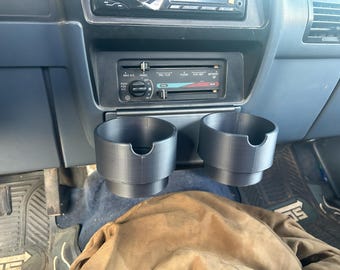 May include: A pair of black, 3D-printed cup holders designed for a vehicle's dashboard. The cup holders are cylindrical with a small notch on the side. The dashboard is blue and has climate control dials and vents.