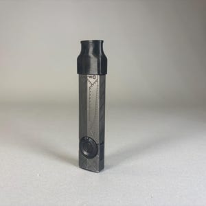 Puede incluir: Un dispositivo rectangular gris oscuro con una parte superior negra. El dispositivo tiene una superficie texturizada con un botón circular y detalles geométricos. La parte superior es cilíndrica. El dispositivo mide aproximadamente 10 cm de alto.