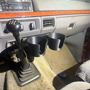 May include: A black dual cup holder attachment mounted in a vehicle interior, positioned next to a manual gear shift lever with a black knob featuring a white shift pattern diagram, set against a gray dashboard with wood grain trim accents.