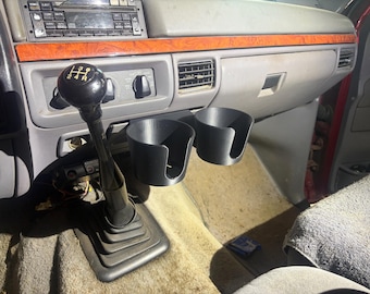 May include: A black dual cup holder attachment mounted in a vehicle interior, positioned next to a manual gear shift lever with a black knob featuring a white shift pattern diagram, set against a grey dashboard with wood grain trim accents.