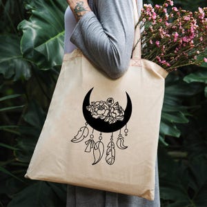 Op de afbeelding: Beige draagtas met een zwart maansikkelontwerp gevuld met bloemen en veren. De tas is gemaakt van een lichtgekleurde stof. De tas is gevuld met roze bloemen. Het ontwerp heeft een minimalistische stijl.