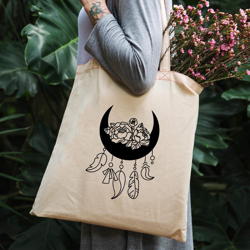 Op de afbeelding: Beige draagtas met een zwart maansikkelontwerp gevuld met bloemen en veren. De tas is gemaakt van een lichtgekleurde stof. De tas is gevuld met roze bloemen. Het ontwerp heeft een minimalistische stijl.