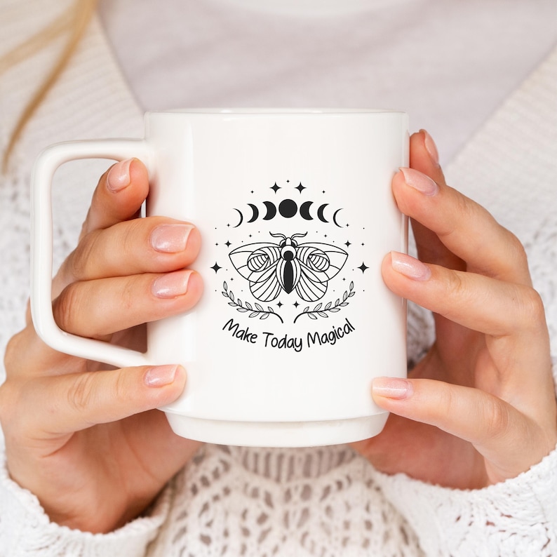 Op de afbeelding: Witte keramische mok met een zwarte illustratie van een mot, maanfasen en sterren. De tekst "Make Today Magical" staat eronder. De mok heeft een groot oor en wordt door een persoon vastgehouden.