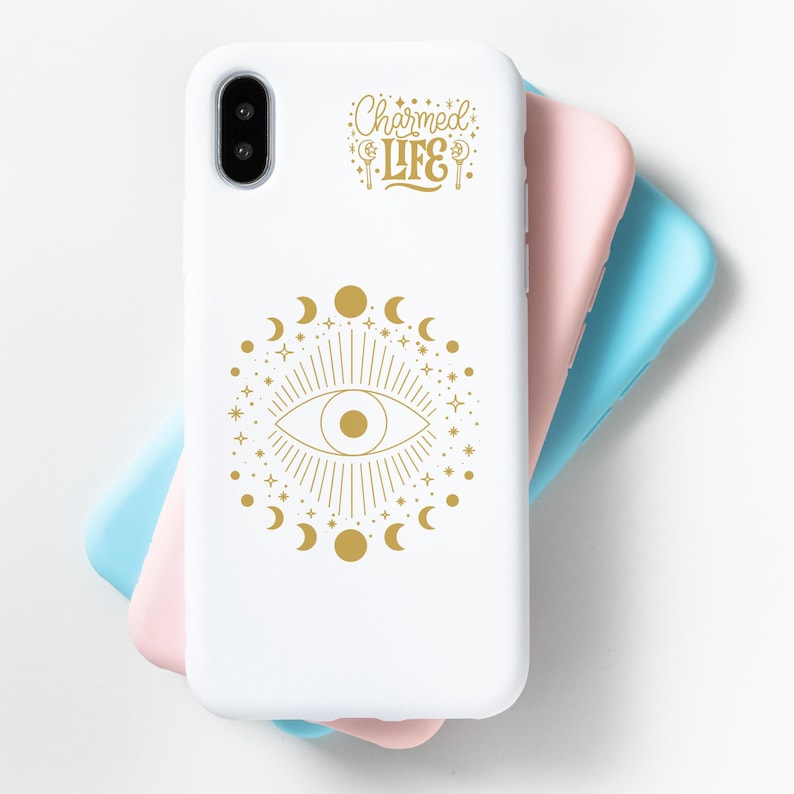 Op de afbeelding: Witte telefoonhoes met een gouden oogontwerp, omringd door manen en sterren, en de tekst "Charmed Life". Een roze en blauwe telefoonhoes is zichtbaar achter de witte hoes.