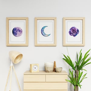 Op de afbeelding: Drie ingelijste kunstdrukken met aquarel illustraties. E&eacute;n toont een paarse maan, een andere een blauwe halve maan en de derde een paarse roos. De frames zijn van licht hout en de prints op een witte achtergrond.