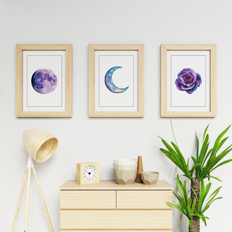 Op de afbeelding: Drie ingelijste kunstdrukken met aquarel illustraties. E&eacute;n toont een paarse maan, een andere een blauwe halve maan en de derde een paarse roos. De frames zijn van licht hout en de prints op een witte achtergrond.