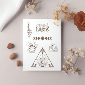 Op de afbeelding: Een witte kaart met bruine illustraties van mystieke bedels, waaronder een hand met een toverstaf, halve manen, een zon, een kristal, een waarzeggerijbal en een driehoek met een oog. De kaart bevat ook de tekst "Mystical Charms".