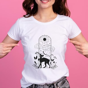 Op de afbeelding: Wit T-shirt met een zwarte afbeelding van een kat in een gewelfd raam, met een zon, wolken en sterren. De kat staat tussen het gebladerte. Het shirt is gemaakt van een zacht materiaal.
