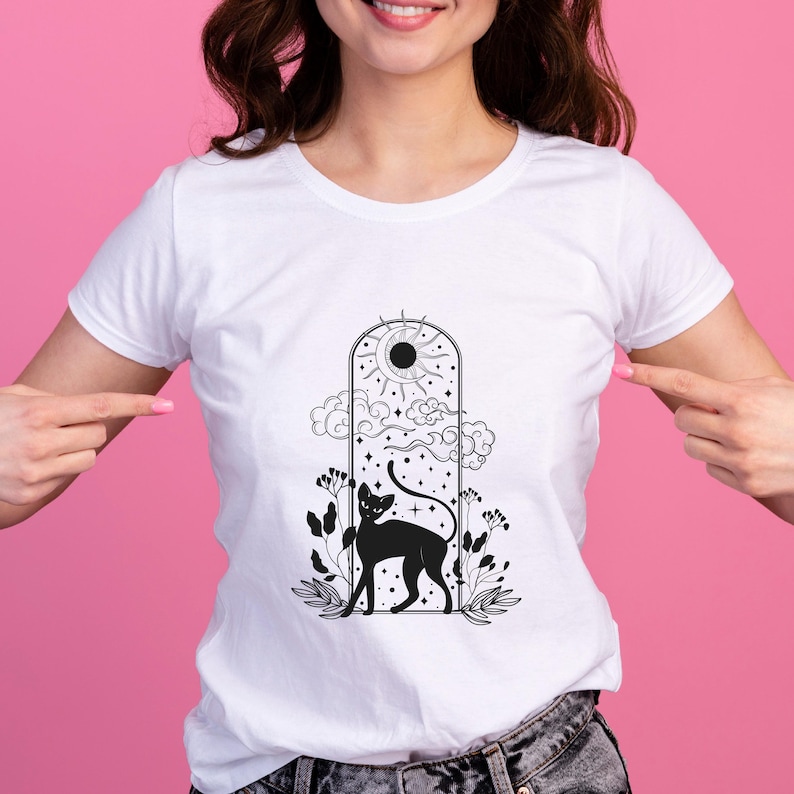 Op de afbeelding: Wit T-shirt met een zwarte afbeelding van een kat in een gewelfd raam, met een zon, wolken en sterren. De kat staat tussen het gebladerte. Het shirt is gemaakt van een zacht materiaal.