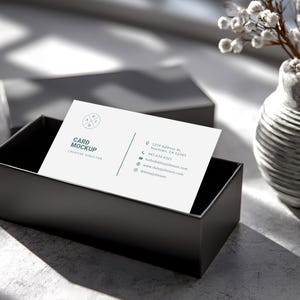 May include: A business card mockup with the text "Card Mockup Creative Director" in a black box. The card has a logo and contact information. A gray vase with white flowers is in the background.