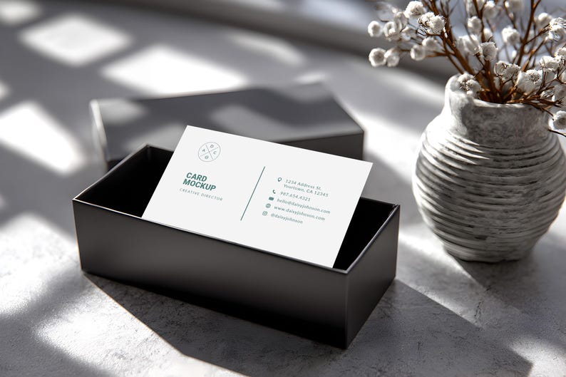 May include: A business card mockup with the text "Card Mockup Creative Director" in a black box. The card has a logo and contact information. A gray vase with white flowers is in the background.