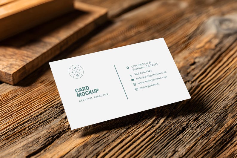 May include: A white business card mockup with the text "CARD MOCKUP" and "CREATIVE DIRECTOR" in a sans-serif font. The card includes contact information and a circular logo. The card rests on a textured wooden surface.