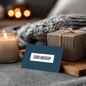 May include: A lit candle in a glass jar, a gift box wrapped in striped paper, and a business card mockup with the text "CARD MOCKUP" on a blue background. The items are arranged on a gray surface with a cozy, blurred background.