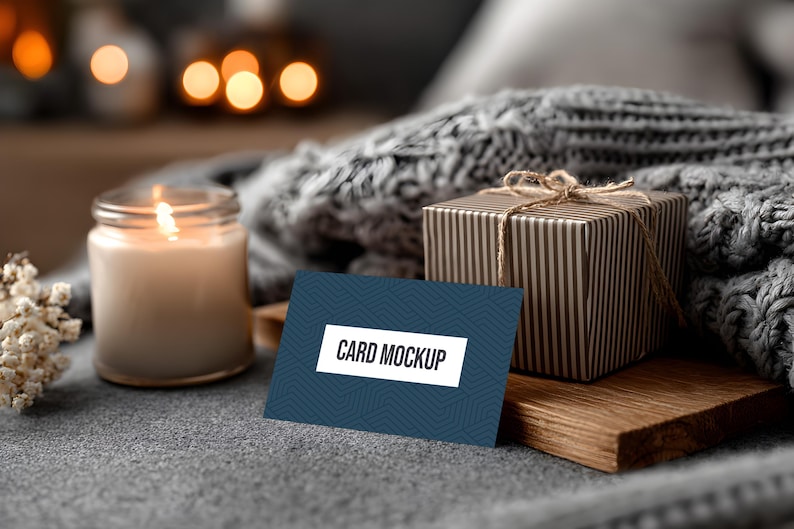 May include: A lit candle in a glass jar, a gift box wrapped in striped paper, and a business card mockup with the text "CARD MOCKUP" on a blue background. The items are arranged on a gray surface with a cozy, blurred background.