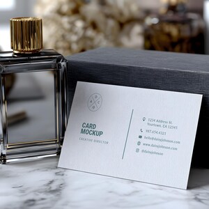May include: A clear glass perfume bottle with a gold cap sits next to a dark gray box and a business card. The business card reads "Card Mockup, Creative Director" with contact information. The perfume bottle has a rectangular shape.