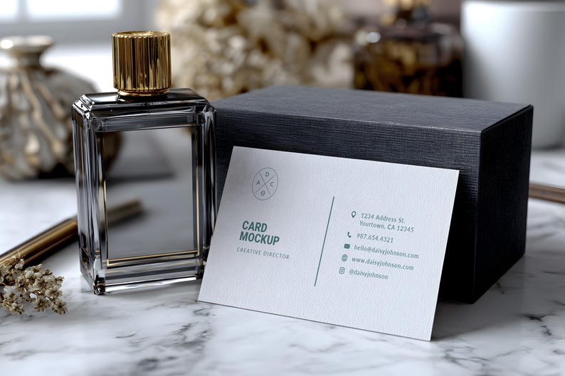 May include: A clear glass perfume bottle with a gold cap sits next to a dark gray box and a business card. The business card reads "Card Mockup, Creative Director" with contact information. The perfume bottle has a rectangular shape.