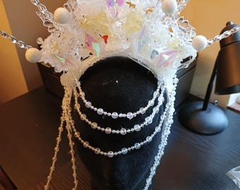 May include: A white crystal crown with a beaded chain. The crown is decorated with white beads and iridescent crystals. The chain is made of clear beads.