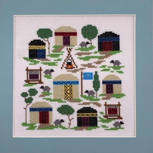May include: Cross-stitch art featuring a village scene with several yurts, trees, and sheep. The yurts have various colors and patterns, with a campfire and flag in the center. The artwork is framed in a white mat and a light blue frame.