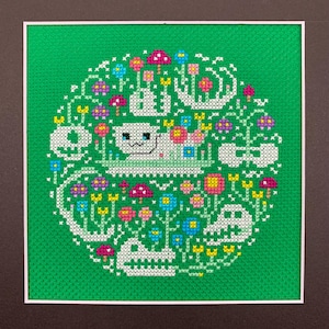 May include: A square framed cross-stitch embroidery on bright green fabric featuring a central white cat surrounded by white animal skulls, colorful pink, yellow, and blue flowers, and small mushrooms arranged in a circular pattern.