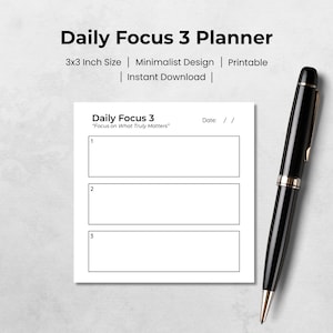 May include: A white Daily Focus 3 planner with three rectangular sections for notes, a date line, and the text "Focus on What Truly Matters." The planner is 3x3 inches in size. A black pen with gold accents rests on the right side.