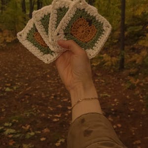 May include: A hand displays four square crochet coasters. Each coaster has a white border, a green centre, and a brown floral design. The background shows a forest with autumn foliage.