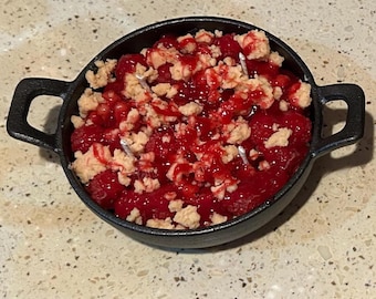 May include: Two individual servings of raspberry crumble in small, black cast iron skillets. The dessert features a vibrant red raspberry filling topped with a golden brown crumble and drizzled with a red sauce. One skillet has a handle, the other has two small handles.