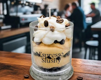 May include: A clear glass candle with a layered design resembling an espresso slushy. The candle features a creamy white top, a light brown middle layer, and a dark layer at the bottom. Coffee beans are scattered on top. The words "Espresso Slushy" are printed on the glass.