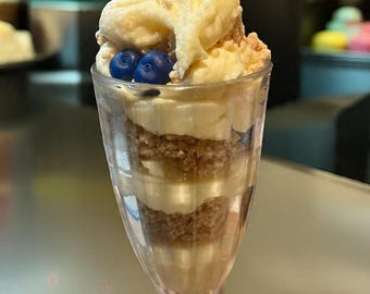 May include: A layered dessert in a clear glass. The dessert features alternating layers of brown and white components, topped with a swirl of cream, blueberries, a lemon slice, and a cinnamon stick. The glass has a decorative base.