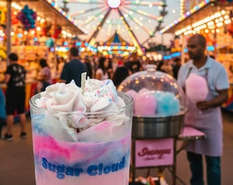 May include: A clear glass candle with a pink base and a whipped cream-like top. The candle has pink, blue, and white layers with the words "Sugar Cloud" in blue. The background shows a carnival with a Ferris wheel and a cotton candy vendor.