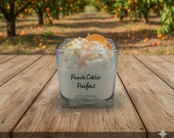May include: A square glass candle with a white, peach-colored, and tan wax design, topped with a peach slice. The candle has the words "Peach Cobler Parfait" printed on the front. The candle sits on a wooden surface.