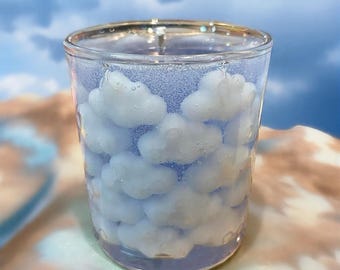 May include: A clear glass candle holder filled with white cloud-shaped wax pieces. The candle holder has a light blue base and is set against a backdrop of a blue sky with clouds. The candle holder is a decorative item.