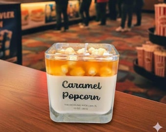 May include: A square glass candle with a caramel popcorn scent. The candle has a white base, a caramel-colored top layer, and is topped with popcorn kernels. The label reads "Caramel Popcorn".
