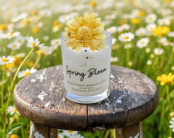 May include: A clear glass candle holder with a white candle and a yellow flower on top. The candle is labeled "Spring Bloom." The candle sits on a small, round, distressed wooden stool. The background is a field of daisies.