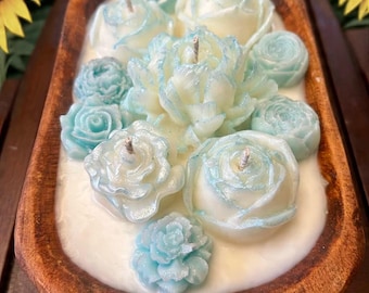 May include: A wooden bowl filled with white wax and several flower-shaped candles. The candles are a mix of light blue and white, with visible wicks. The bowl has a rustic, natural wood finish.