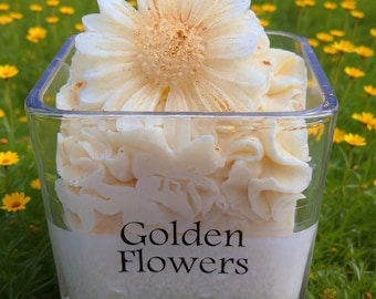 May include: A square glass candle holder with a white label that reads "Golden Flowers." The candle is topped with a large, cream-colored flower and surrounded by a field of yellow flowers. The candle is labeled as a hand-poured soy candle, 10 oz.